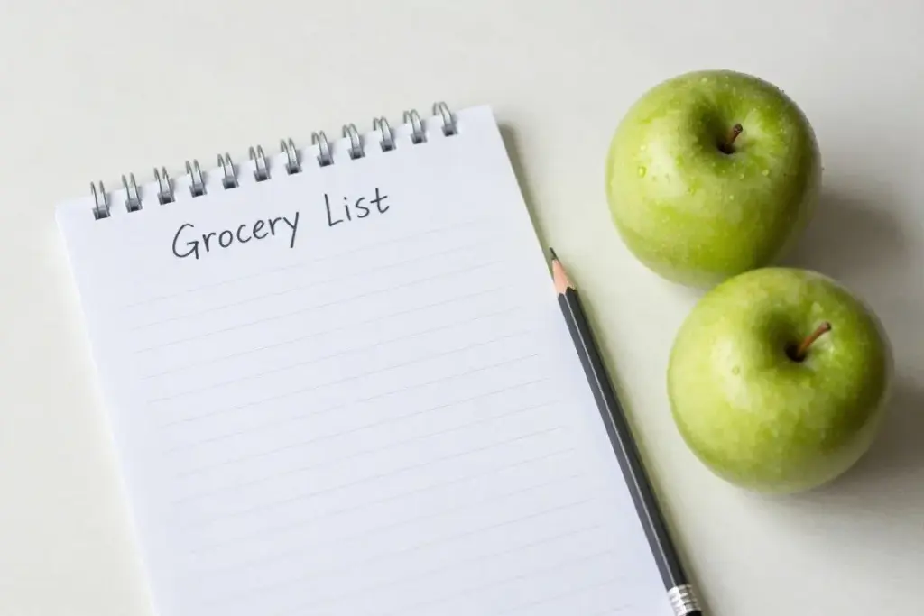 A handwritten shopping list and recipe planner lying next to fresh apples and a wooden pencil