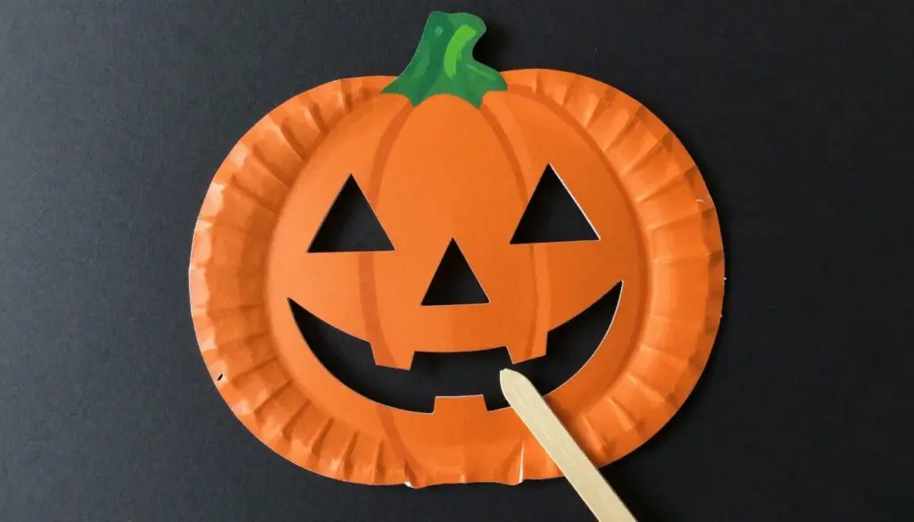 A paper plate jack o lantern mask with cut-out eyes and a craft stick handle